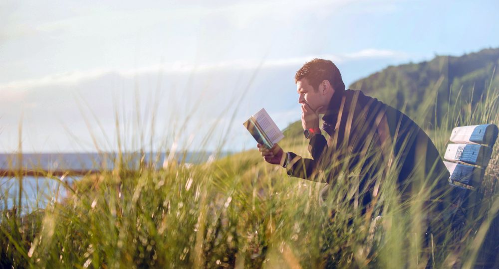 A man reads a book while sitting on a bench in tall grass near the water.
